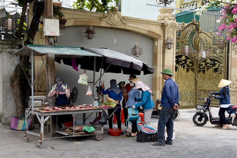 Hong holds up the peace sign while standing behind her stall. Six customers are in a queue to her left. One of them also holds up the peace sign. All are wearing hats and/or face coverings.